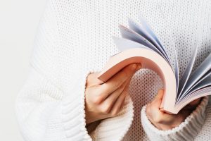 Close view of woman in white woolen sweater holding an open book with pink cover in hands. Long wide banner with free space for your mock up of reading book concept background.