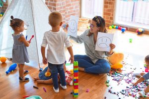 Beautiful psychologist and group of toddlers make therapy using emotions emojis around lots of toys at kindergarten